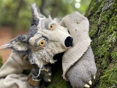 Eine Pupenfigur aus Stoff, ein grauer Wolf, lehnt sich  an einen Baum im Wald.